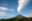 Volcano with smoke coming out of top and up into blue sky dotted with floating clouds.