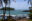 Coast dotted with rocks with palm trees has gentle waves rolling in while beyond is a round island covered in palm trees