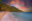 Wide sandy beach with waves gently lapping at shoreline. Beyond are palmtrees and forested mountains under a fiery red sky dotted with clouds. People are swimming just out from shore and colourful painted boats are moored beyond