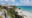 Visitors on a viewing platform overlook a rocky, palm-fringed shore, where boulders meet white sand and turquoise sea, a square stone structure rising behind them.