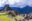 An overview of stone buildings without roofs and stone walls sitting across grassy mountain-top with jagged mountains and low clouds in the background