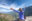 Woman wearing purple jacket and khaki hat, her arms raised in the air, sitting on a rocky ledge overlooking mountains